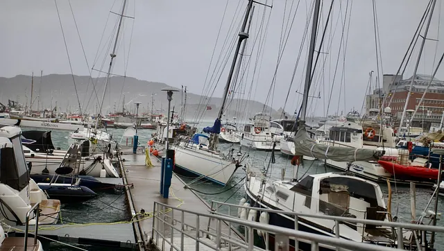 Seilbåt fortøyd ved brygge i regnvær med andre båter og havn i bakgrunnen. ⛵
