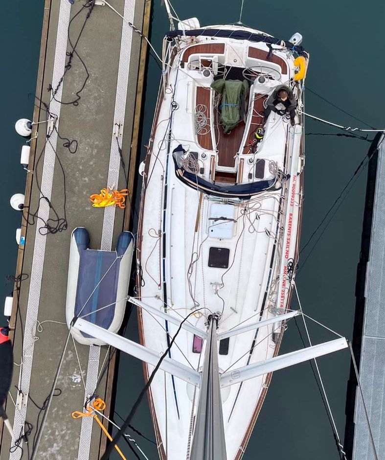 Seilbåt fortøyd mellom brygger sett ovenfra, med tauverk og utstyr synlig på dekk.