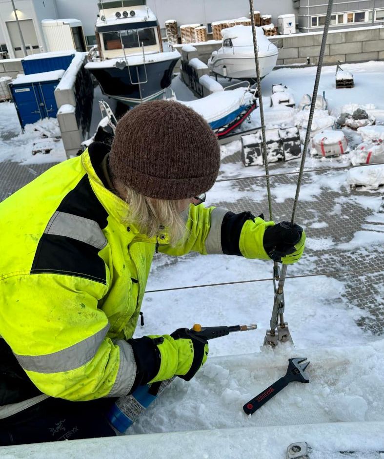 Person i refleksjakke jobber med rigg og vaier på snødekt seilbåt ved opplagsplass.